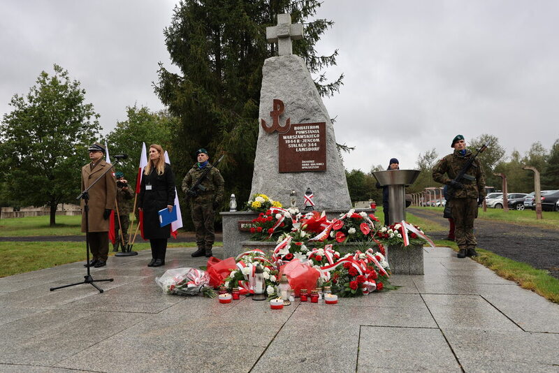 Zdjęcie przedstawia pomnik pod którym złożone są kwiaty, po lewej i po prawej stronie stoi żołnierz