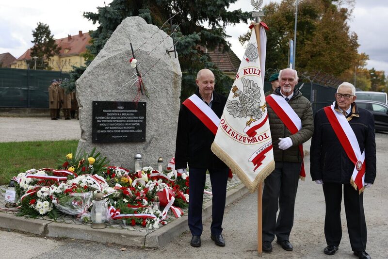 na zdjęciu obelisk z kwiatami, stojący przy nim poczet sztandarowy