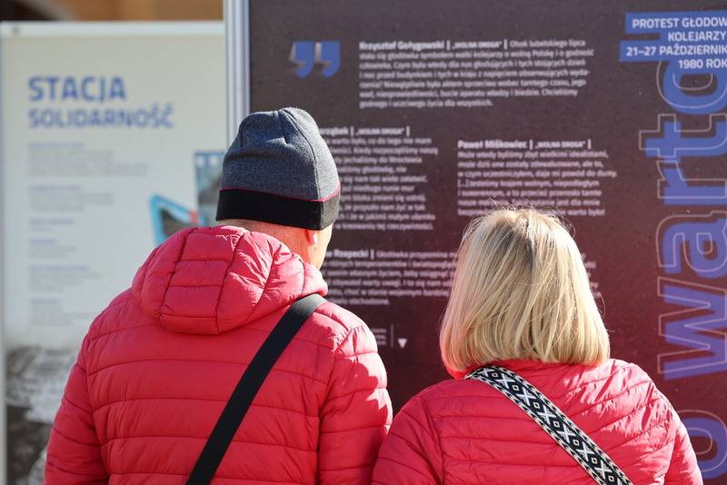 ludzie stojący przed planszą i oglądający wystawę