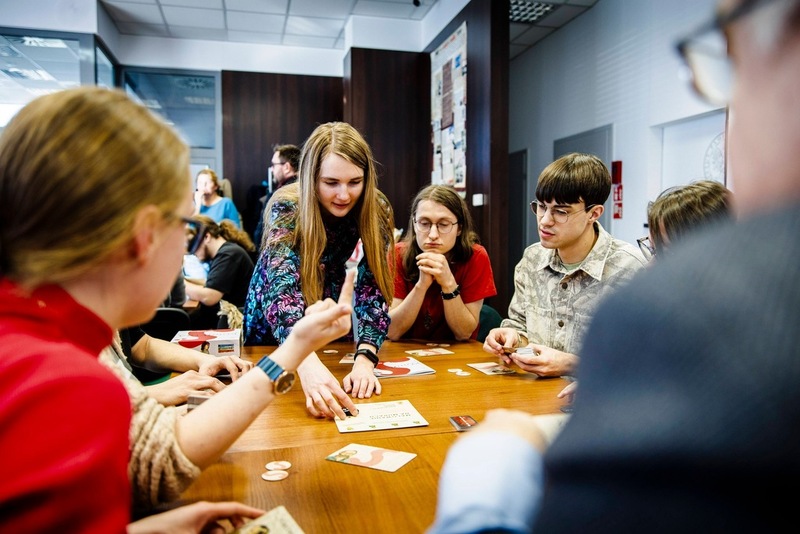 zdjęcie przedstawia osoby siedzące przy stole i grające w grę, nad nimi pochyla się instruktor