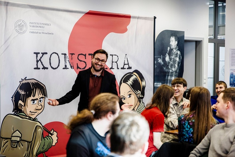 zdjęcie przedstawia osoby siedzące przy stole i grające w grę, przemawia do nich Grzegorz Kowal autor gry