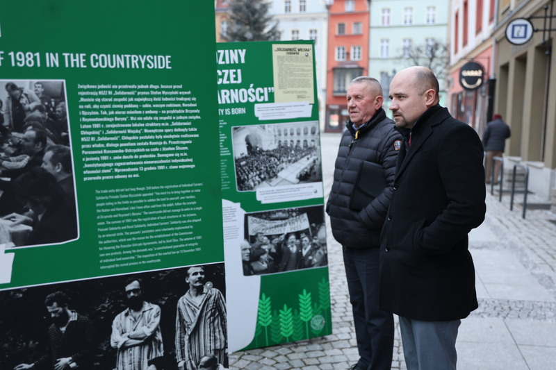 zdjęcie przedstawia wystawę stoją na rynku, przy planszach dwóch obserwujących