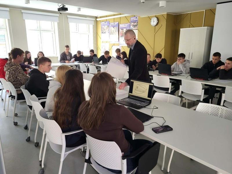 Widok na salę, na stole stoją komputery, gdzie młodzież realizuje zadania. Na środku prowadzący zajęcia Artur Cieślik