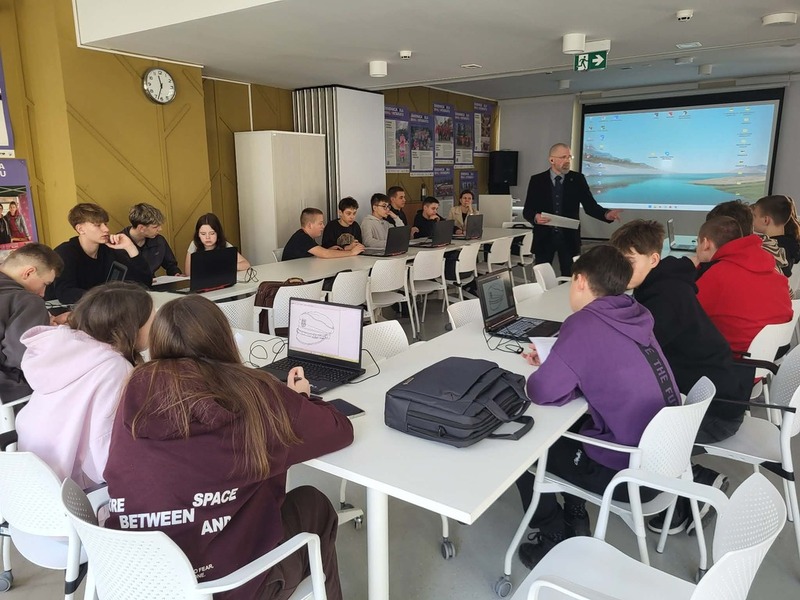 Widok na salę, na stole stoją komputery, gdzie młodzież realizuje zadania. Na środku prowadzący zajęcia Artur Cieślik