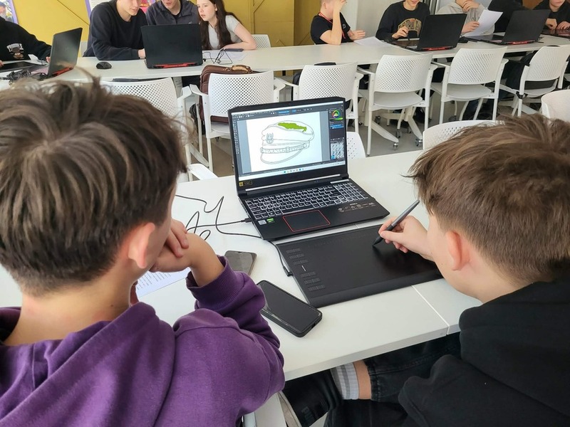 Widok na salę, na stole stoją komputery, gdzie młodzież realizuje zadania. Na środku prowadzący zajęcia Artur Cieślik