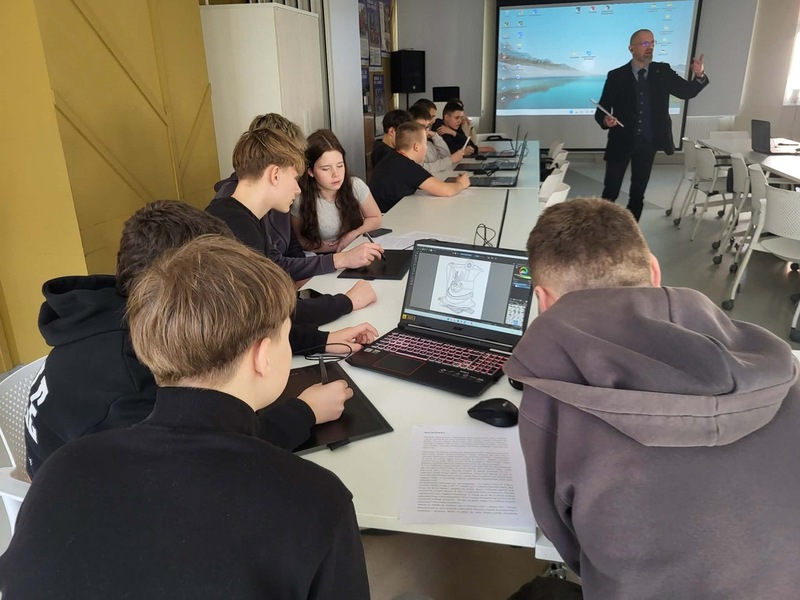 Widok na salę, na stole stoją komputery, gdzie młodzież realizuje zadania. Na środku prowadzący zajęcia Artur Cieślik