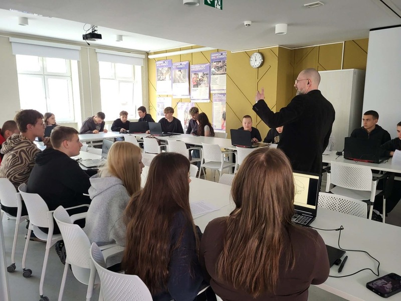 Widok na salę, na stole stoją komputery, gdzie młodzież realizuje zadania. Na środku prowadzący zajęcia Artur Cieślik